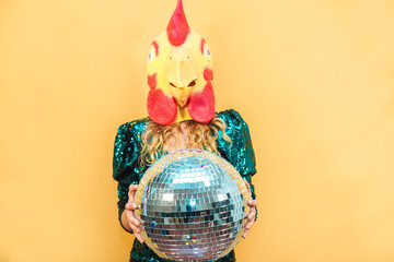 Wall Mural - Young woman wearing chicken mask while holding disco ball at new year's party - Soft focus on face