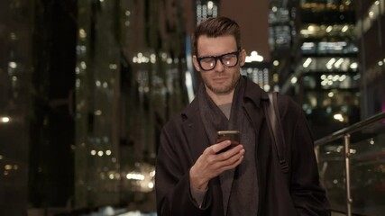 Fototapete - Businessman in eyeglasses with phone in hand on background of buildings with lights on. Gimbal night shot of caucasian man in black coat texting in phone near business building
