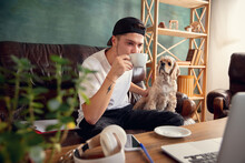 Coffee Break. Young Man Sitting On Brown Sofa With His Cute Dog, Pet American Cocker Spaniel During Coronavirus Or Covid-19 Quarantine. Cozy Office Workplace, Remote Work, E-learning Concept.