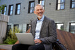© Kostiantyn - Ideas for business. Portrait of cheerful elegant middle aged businessman smiling while working on his laptop, sitting on the bench near modern building outdoors