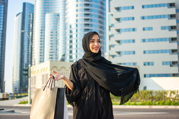 Wall Mural - Beautiful young arab muslim woman in abaya clothes holding shopping bags and walking on the city street. Shopping time. Modern skyscrapers in the background.