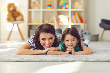 Happy mother and smiling daughter lying together on carpet on floor and looking at camera