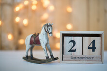 Cube Wooden Calendar Showing Date On 24th December With Old Toy Rocking Horse Over Bokeh Background. Advent Calendar, Christmas Background, Copy Space