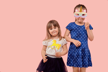 Two little cute girls sisters are celebrating their birthday, holding a cardboard unicorn. Pink background.