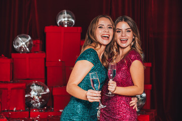 Two cheerful happy girls having a new year party, celebrating christmas. They drink champagne and open presents. They wear sexy dresses that curves their body in sexy way. Many red presents on back.