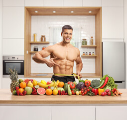 Sticker - Muscular man in a kitchen measuring waist and posing with a pile of healthy fruits and vegetables