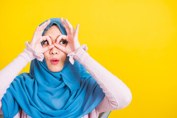 Wall Mural - Asian Muslim Arab, Portrait of happy beautiful young woman Islam religious wear veil hijab funny smile she show gesture fingers in okay gesture symbol, OK sign over her eyes isolated yellow background