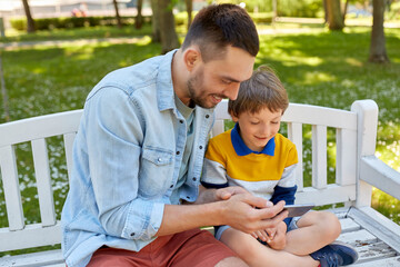 Wall Mural - family, fatherhood and technology concept - happy father and little son with smartphone sitting on bench at summer park