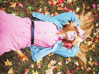 Wall Mural - Red-haired cheerful girl lies in a meadow covered with maple leaves with her palms folded above her head