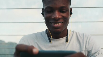 Poster - A happy african american man with earphones is watching a video on his smartphone sitting outside on the city bridge