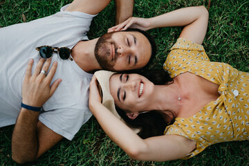 Wall Mural - A brunette girl in a dress and a hat is lying near her boyfriend with a beard on the grass in the park in Valencia. A couple of tourists with closed eyes is enjoying the summer in Spain.