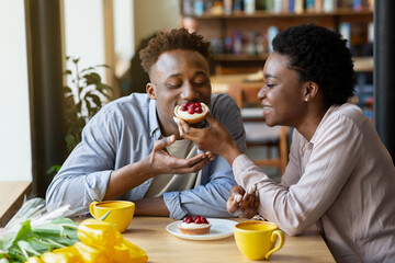 Lovely black woman feeding tasty berry tartlet to her boyfriend at cozy city cafe