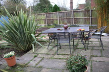 Landscape Of Home Garden In Winter Environment With Metal Table Chairs On Stone Patio, Lantern & Apple Windfalls Background Of Wood Fencing Brick Path Soil Plants And Blue Shed In England On Grey Day