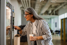 Happy White-haired Woman Using Vending Machine While Drinking Coffee