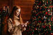 © dikushin - Side view of serious redhead young woman typing messages on mobile phone near Christmas tree. Middle shot portrait of female chatting online on cellphone at cozy living room with festive interior