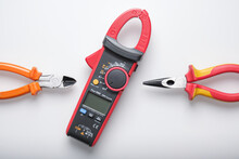 Red Digital Clamp Meter Placed On Center With Orange Wire Cutter And Red Pliers Placed On The Sides Isolated On White Background. Electrical Engineering Tools Composition.