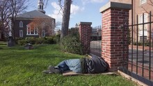 A Young Caucasian Homeless Man Is Sleeping On Grass In The Graveyard In Front Of  A Church In Downtown Alexandria. Man Is Wearing Jeans And Sweatshirt
