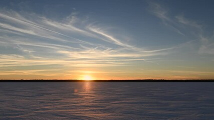 Wall Mural - Beautiful sunset time lapse over a snow-covered lake with silhouettes of trees in the distance. The sky is blue, yellow, and orange with wispy clouds and the occasional jet vapor trail. 
