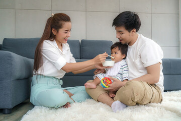 Asian mother feeding his 6 months old baby boy with solid food with spoon and father sitting near to cheer her son to eat food in living room at home..