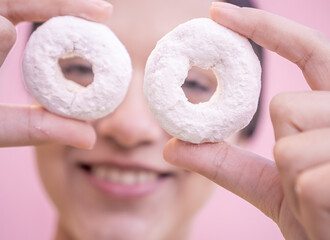 Latin woman holding two delicious donuts in front of her eyes with a pink background.