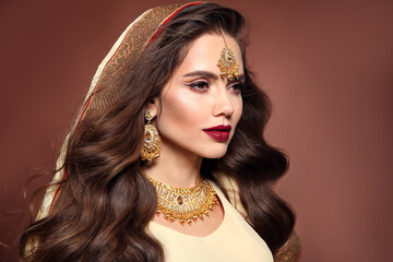 Wall Mural - Beautiful woman with long wavy hair style and bright makeup. Young hindu brunette model with kundan golden jewelry set isolated on brown studio background.