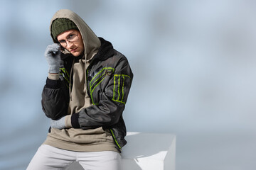 young trendy man in hat, gloves and anorak sitting on white cube while adjusting glasses on grey