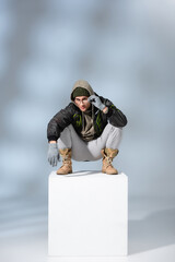 stylish man in hat, gloves and parka adjusting glasses while sitting on white cube on grey