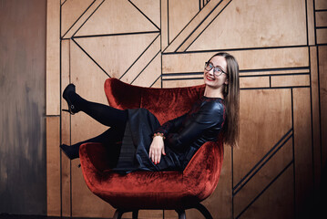 Young brunette woman, wearing fancy black leather dress and eyeglasses, sitting on red velvet Santa armchair in front of wooden wall, Merry Christmas and Happy New year celebration. Female portrait