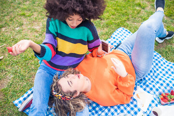 two multiethnic female best friends having picnic in a park having fun celebrating using smartphone laughing enjoying life