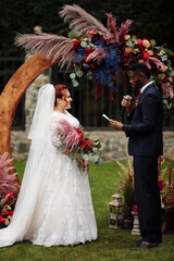 Handsome African American man with beautiful white woman stand near decorated arch, adorable couple on wedding ceremony, attractive groom say oath to lovely bride, marriage concept