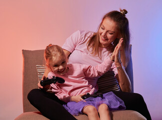 Excited young mother and cute little daughter sit on couch holding gamepad controller while playing video games at home together, have fun in in living room with red blue neon light