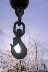 A crane hook hangs against a backdrop of a blue sky with the sun shining through the hook.