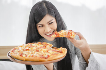 Beautiful happy smiling girl sitting on the bed showing a whole pizza and trying to eat a piece of pizza in her hand for her breakfast