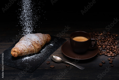 Tela on the rustic wooden table and dark background, a cup of creamy espresso with croissants and cascade of powdered sugar