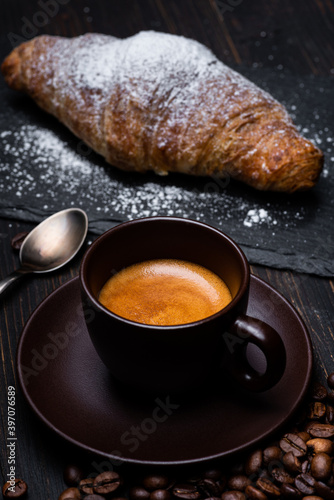 Tela on the dark rustic wooden table, a creamy cup of Italian coffee in the foreground and a croissant sprinkled with powdered sugar