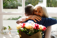 Senior Caucasian Woman With Flower Bouquet Hugging Her Husband In The Kitchen At Home