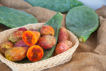 Group of prickly pears on a piece of jute, green leaves and a wicker basket full of fruits