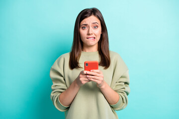 Wall Mural - photo portrait of anxious woman biting lower lip holding phone in two hands isolated on vivid teal c