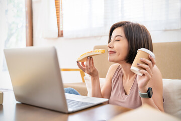 ortrait of happy asian woman working on laptop computer, have lunch break eating pizza in sofa living room using internet. Delivery, work from home quarantine new normal with copy space background