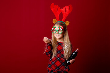 Wall Mural - a little girl in Christmas glasses holding a bottle with a drink. Christmas concept, red background, text space