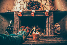 Feet In Woollen Socks By The Christmas Fireplace. Woman Relaxes By Warm Fire With A Cup Of Hot Drink And Warming Up Her Feet In Woollen Socks. Close Up On Feet. Winter And Christmas Holidays Concept.