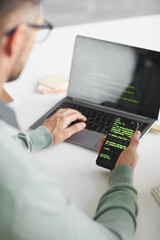 Wall Mural - Rear view of man sitting at the table and programming software on laptop and mobile phone