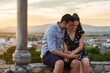 © Cristian Blazquez/ADDICTIVE STOCK - Gentle couple sitting on stone border and embracing while watching video together on cellphone on background of old city under sunset sky