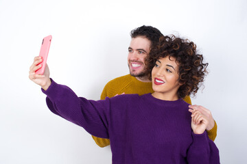 Wall Mural - Portrait of a Young couple wearing knitted sweater standing against white wall  taking a selfie to send it to friends and followers or post it on his social media.