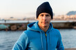 © Galdric Penarroja/ADDICTIVE STOCK - Confident adult ethnic male in warm sportive hoodie and knitted hat looking at camera while standing on embankment near sea at sunset time