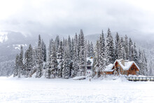 Picturesque Winter Landscape Of Snowy Valley With Wooden House Surrounded By Coniferous Woods And Rocky Mountains Under Cloudy Sky