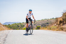 Full Length Of Active Female Bicyclist In Helmet And Sportswear Riding Bike On Asphalt Road Near Mountain Ridge In Summer Day