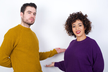 Wall Mural - Portrait of Young couple wearing knitted sweater standing against white wall  with arm out in a welcoming gesture.