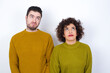 © Jihan - Young couple wearing knitted sweater standing against white wall has worried face looking up lips together, being upset thinking about something important, keeps hands down.