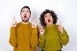 © Jihan - Young couple wearing knitted sweater standing against white wall being amazed and surprised looking and pointing up with fingers showing something strange.